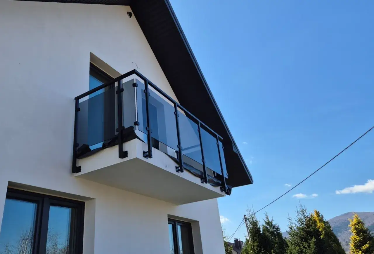 Nowoczesny balkon z czarnymi, szklanymi barierkami, idealnie komponujący się z białą elewacją domu.