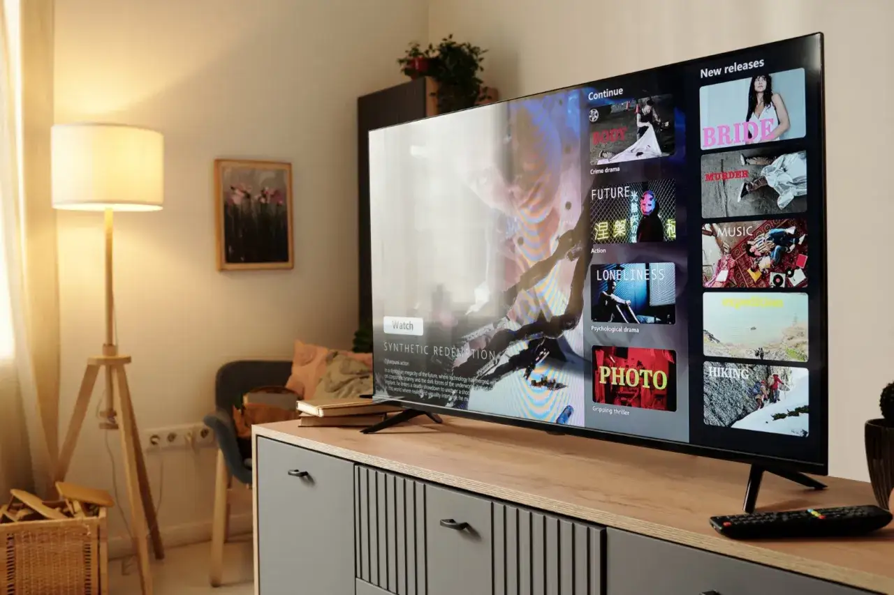Telewizor 55 cali wyświetla menu z filmami. Obok stoi lampa, a na szafce książki.