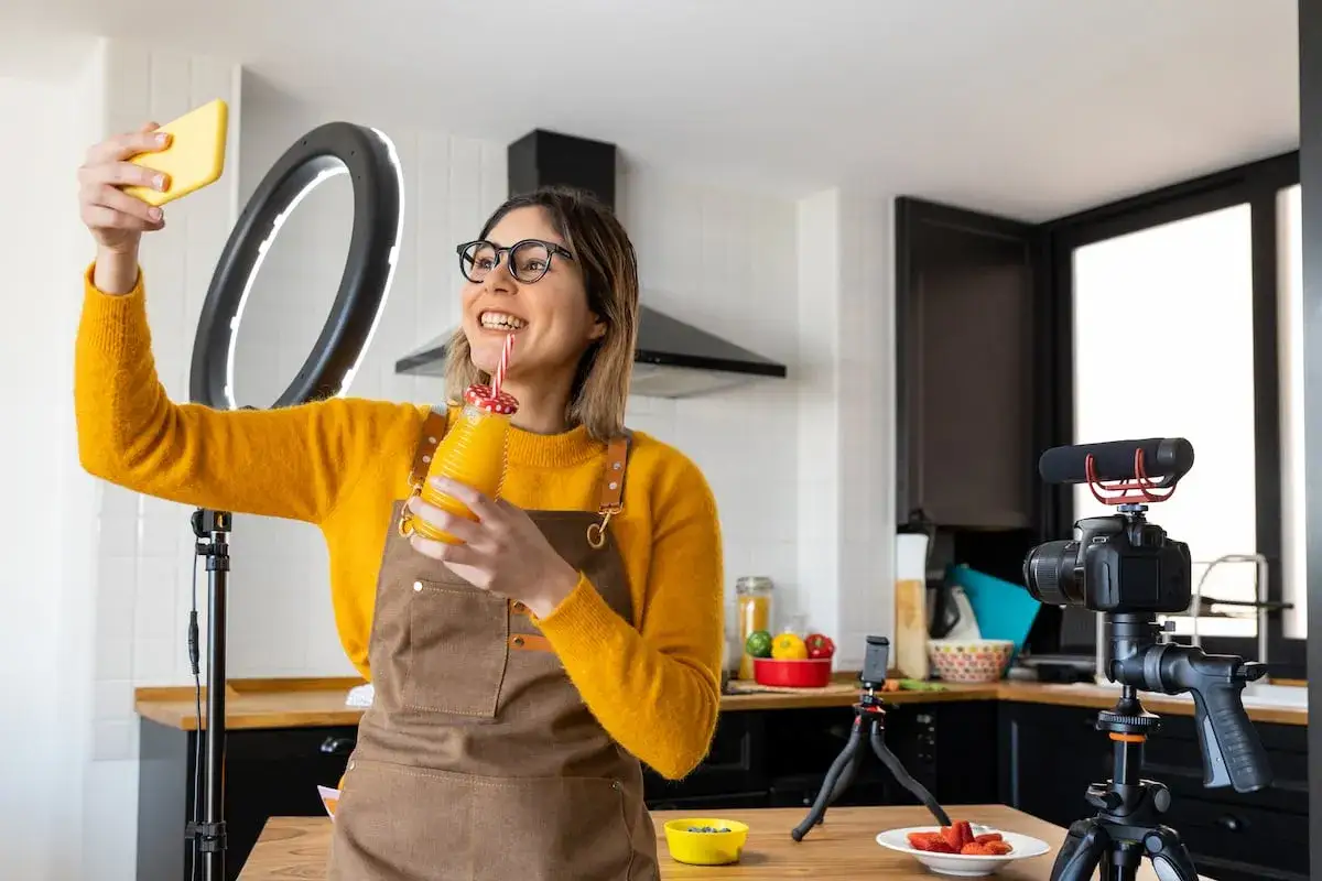 Mujer sonriente con delantal graba un video para aprender como ser influencer, con un aro de luz y c&aacute;mara profesional en su cocina.
