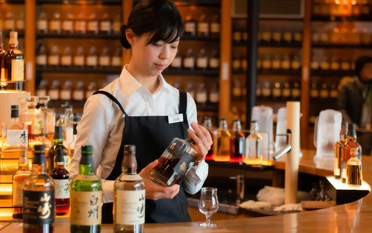 Eine Kellnerin präsentiert eine Flasche mit gutem Scotch. Regale voller Whiskyflaschen im Hintergrund.