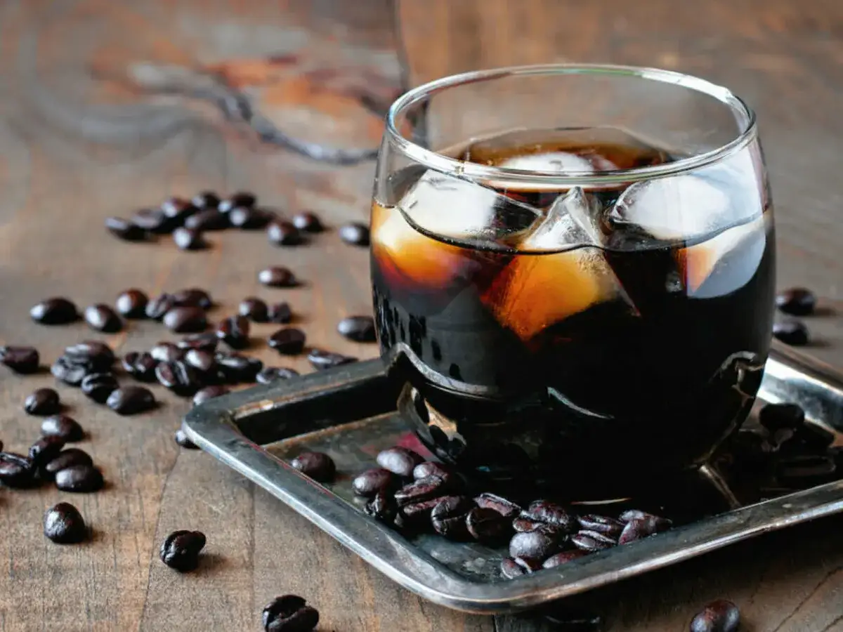 Ett glas med en Black Russian, fylld med is och m&ouml;rkt kaffe, omgivet av kaffeb&ouml;nor p&aring; ett tr&auml;bord.