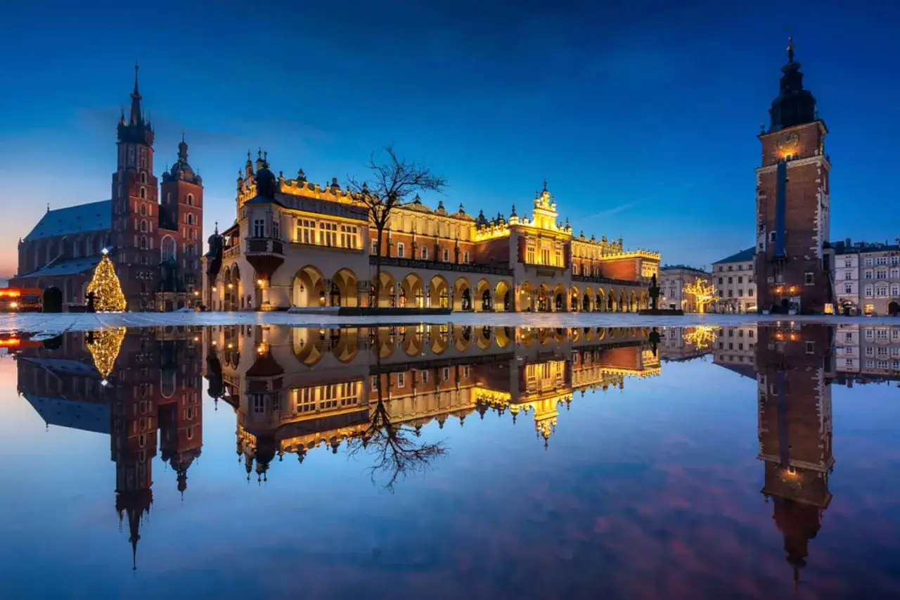 Krak&oacute;w nocą: Rynek Gł&oacute;wny z Sukiennicami, Kościołem Mariackim i wieżą Ratuszową, odbijające się w kałuży. Idealne miejsce na wieczorne atrakcje w Krakowie dla dorosłych.