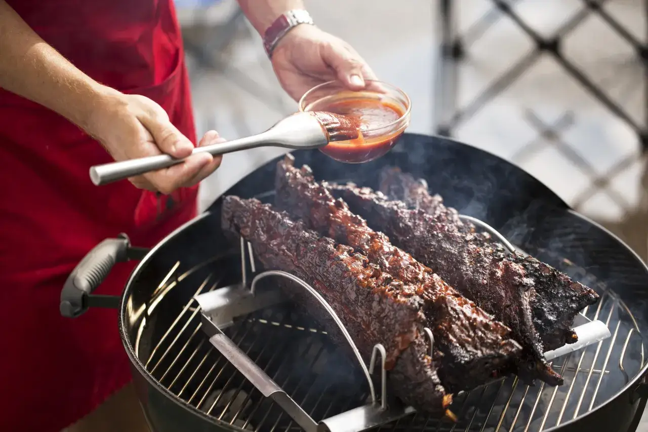 Jak przygotować żeberka na grilla: polewanie ich sosem BBQ z miseczki. Dym unosi się z rusztu.