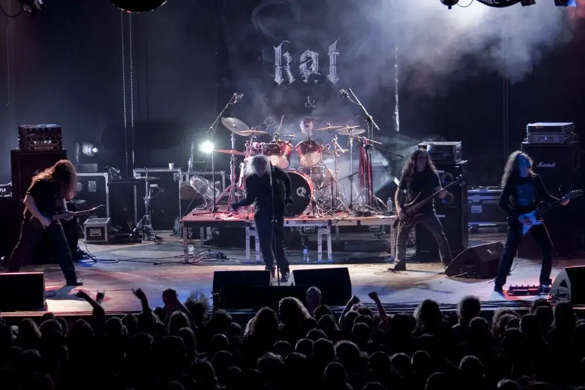 Kat Roman Kostrzewski na scenie z zespołem, grając koncert dla tłumu fanów.