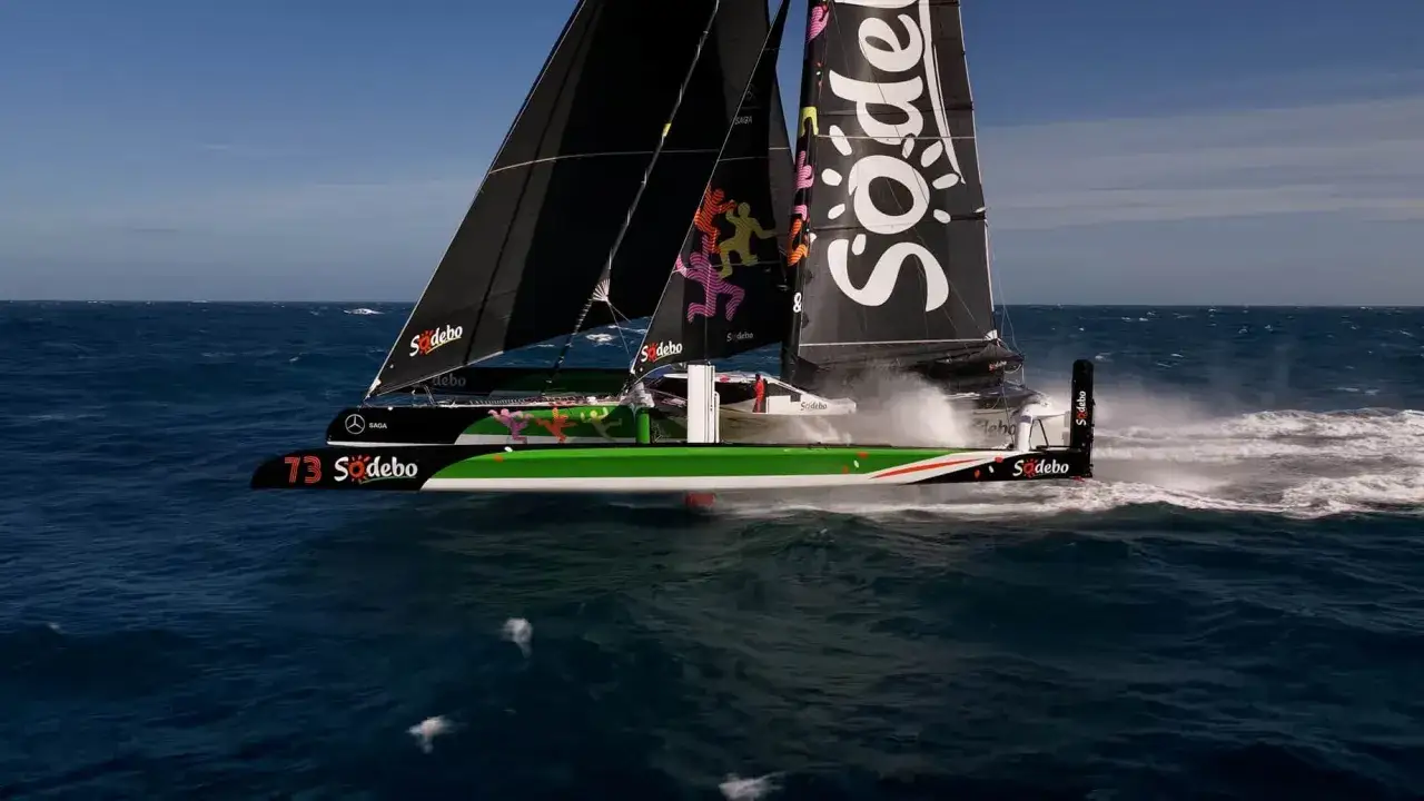 Le trimaran Sodebo Ultim 4 fend les vagues, ses voiles noires portant le logo Sodebo. L'eau éclabousse le pont lors de cette course intense.