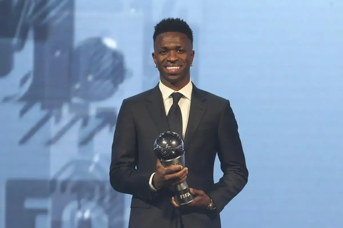 Vin&iacute;cius J&uacute;nior, sonriendo, sostiene un trofeo FIFA. Su atuendo elegante y el premio sugieren que es un jugador excepcional, quiz&aacute;s el MVP en f&uacute;tbol.