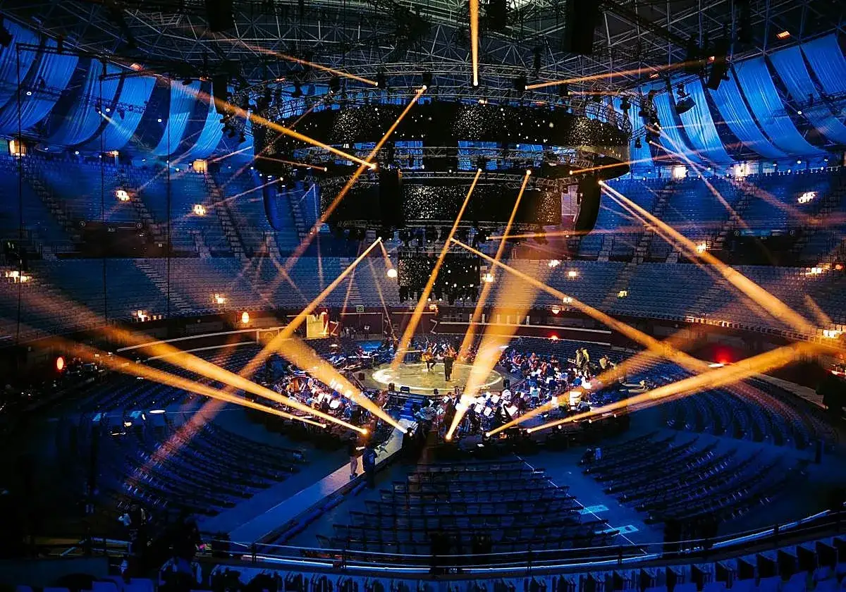 Un concierto con luces naranjas cruzándose sobre un escenario circular. La orquesta y el público están sentados, pero el **bad sound system** arruina la experiencia.