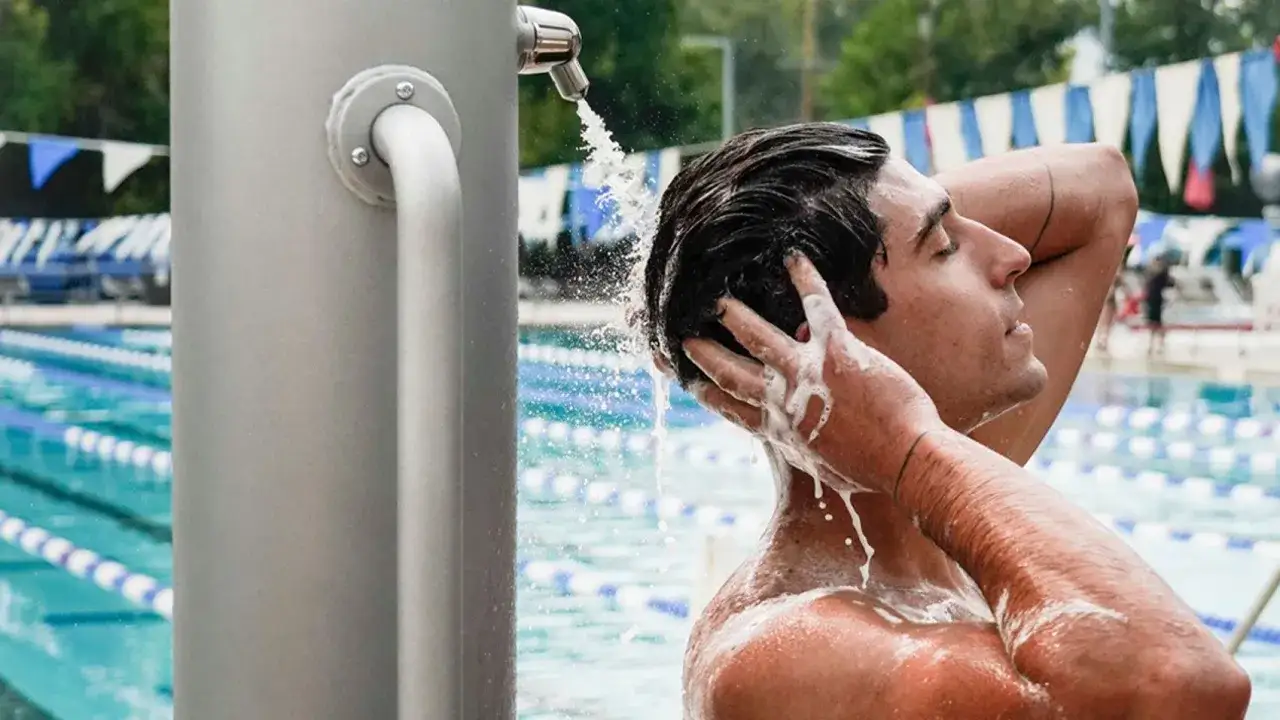 Hombre se enjuaga el pelo oscuro bajo una ducha exterior, con el agua cayendo como si fuera pelo verde piscina.