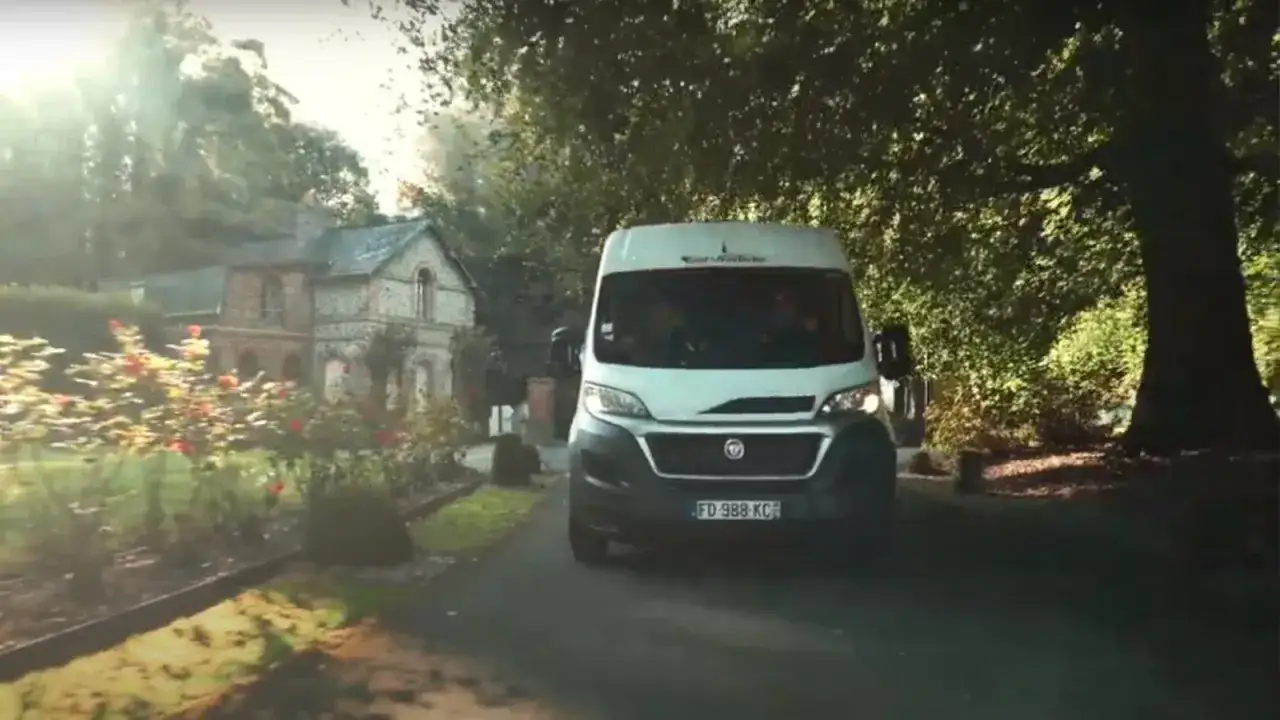 Un spot de Normandie : un camping-car blanc roule sur une all&eacute;e bord&eacute;e de roses, menant &agrave; une maison en pierre.