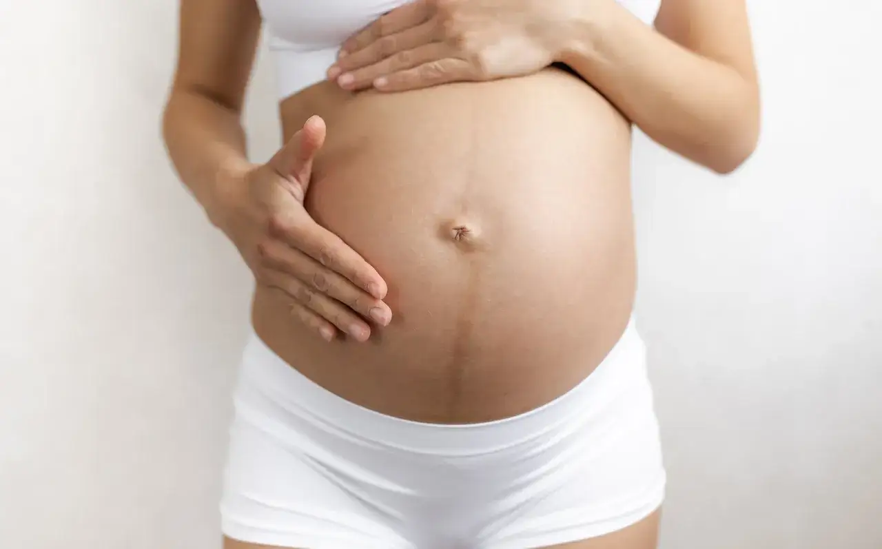 Brzuch kobiety w ciąży z widoczną ciemną linią linea negra. Dłonie delikatnie dotykają zaokrąglonego brzucha.