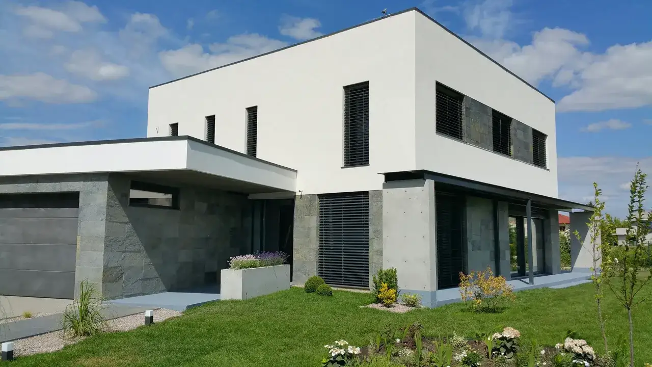 Modernt hus med vit, putsad fasad och stenbeklädd entré. Grönskande trädgård framför.