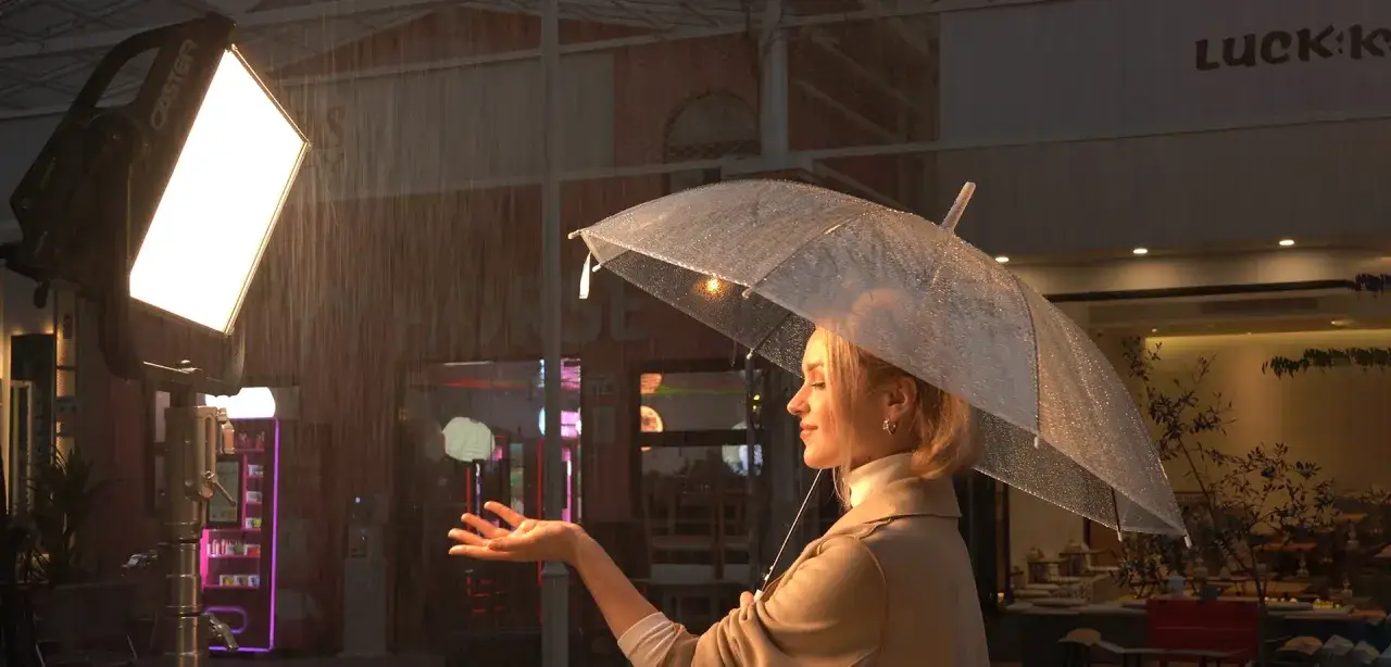 Una donna con un ombrello trasparente sotto la pioggia, illuminata da luci soffuse. La sua mano tesa cattura le gocce d'acqua.
