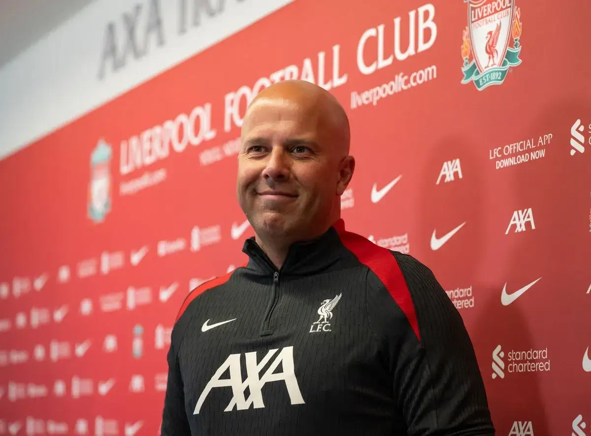Uśmiechnięty mężczyzna w czarnej bluzie z herbem Liverpoolu. Tło z logo klubu i hasłem
