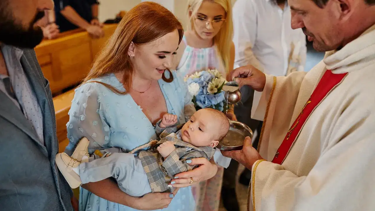 Mama trzyma dziecko do chrztu. Ksiądz polewa mu głowę wodą, pokazując, jak przebiega chrzest.