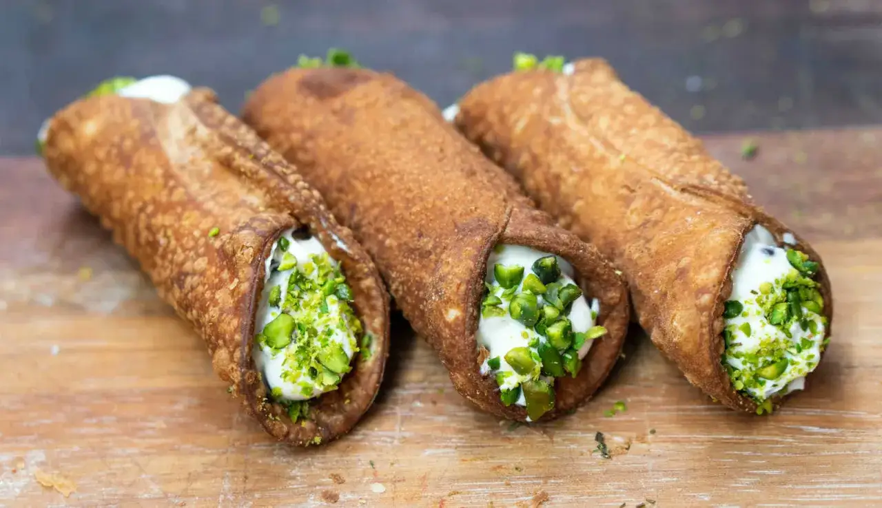 Trzy chrupiące włoskie słodycze, cannoli, wypełnione kremem i posypane pistacjami, leżą na drewnianej desce.