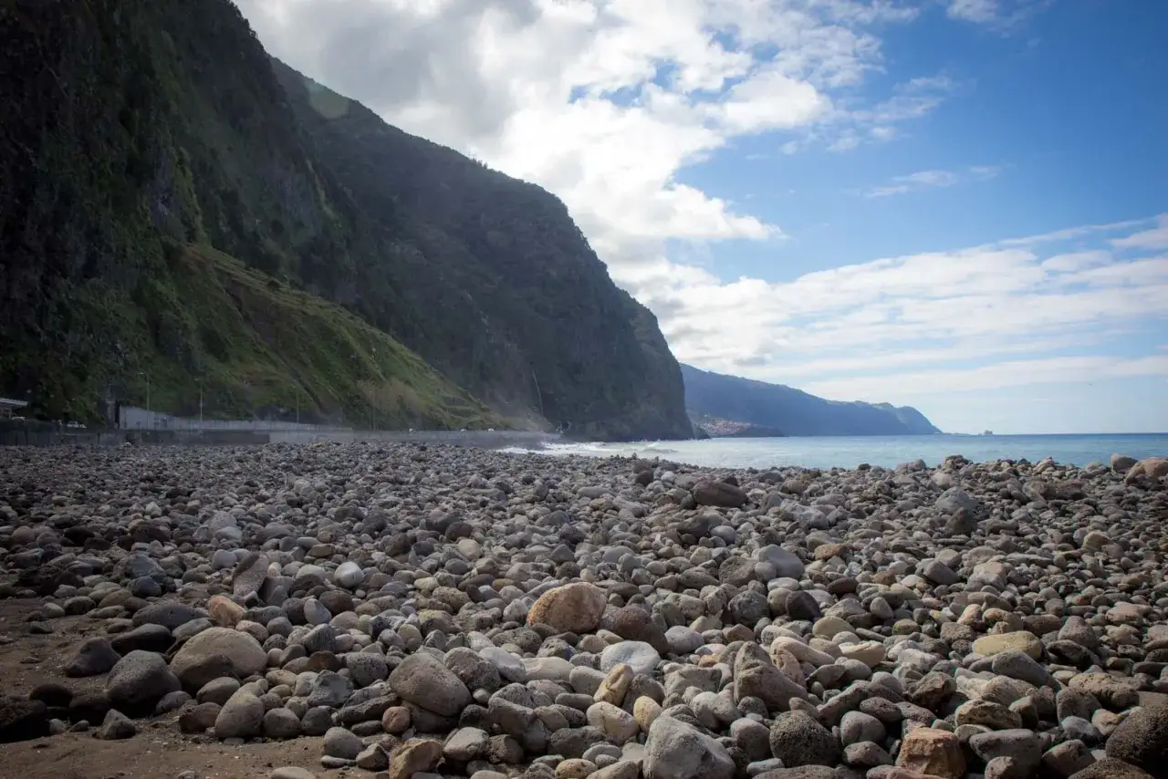 Steiler Berghang trifft auf steinigen Strand am Ribeira da Janela. Das Meer glitzert im Hintergrund.