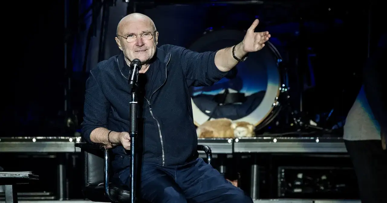 Phil Collins, siedzący na scenie, opowiada o swoich żonach. W tle widoczna jest perkusja.