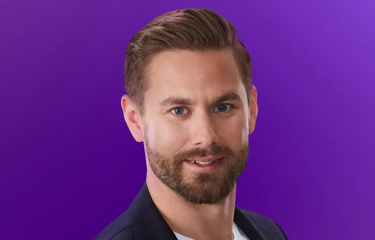 Oskar från Love is Blind, med blå ögon och skägg, ler mot kameran mot en lila bakgrund.