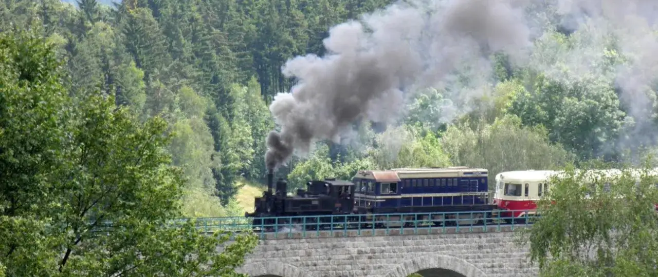 Parowóz z wagonami na kamiennym wiadukcie, dymiący na tle zielonych gór. Idealny na wycieczkę, gdy kupisz bilety na kolej izerską.