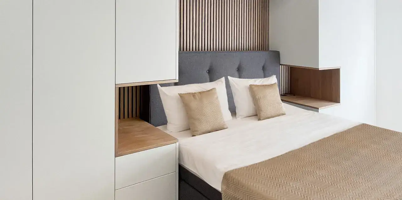 Ein gem&uuml;tliches kleines Schlafzimmer einrichten mit einem Bett, Kissen und einer Wand aus Holzlamellen. Wei&szlig;e Schr&auml;nke und Holzdetails schaffen ein modernes Ambiente.