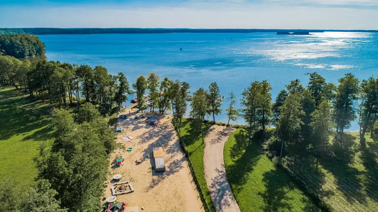 Malowniczy widok na jezioro z plażą i drzewami. W oddali widać żaglówki. Idealne miejsce na wypoczynek w camp park Sonata.
