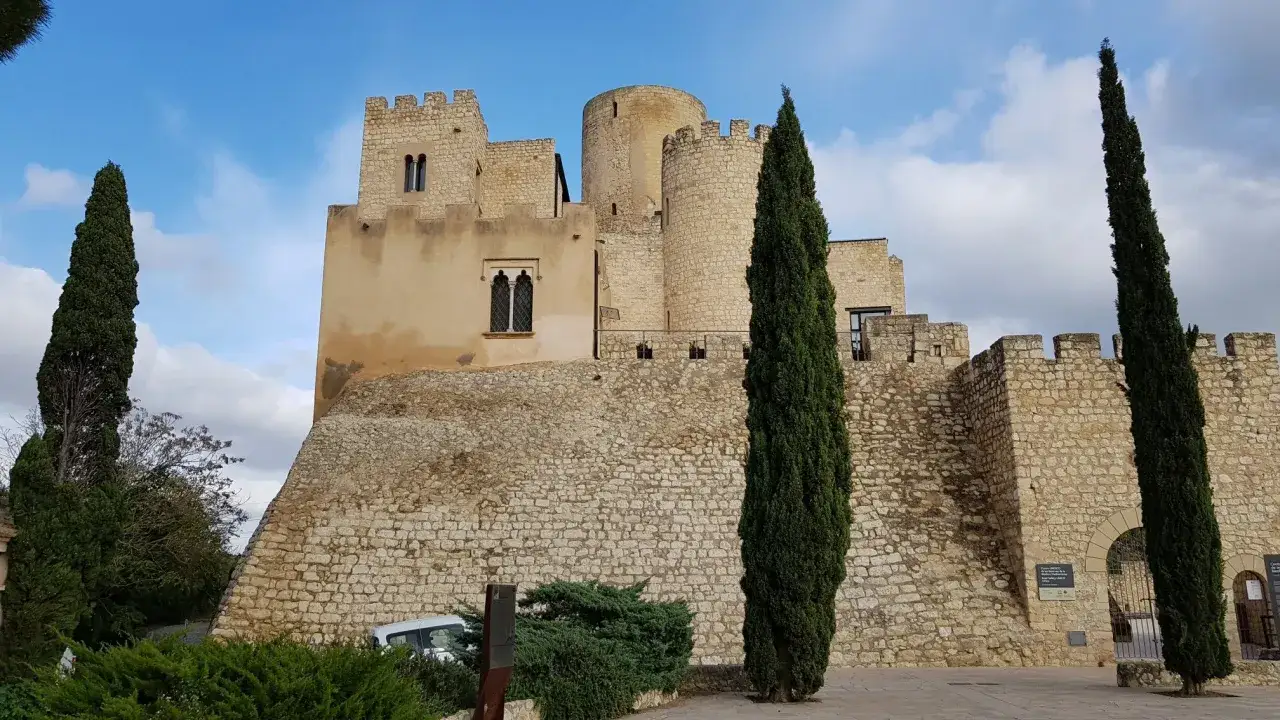 El imponente castillo del Parc del Foix se alza sobre una colina rocosa, flanqueado por cipreses altos y esbeltos bajo un cielo azul con nubes.