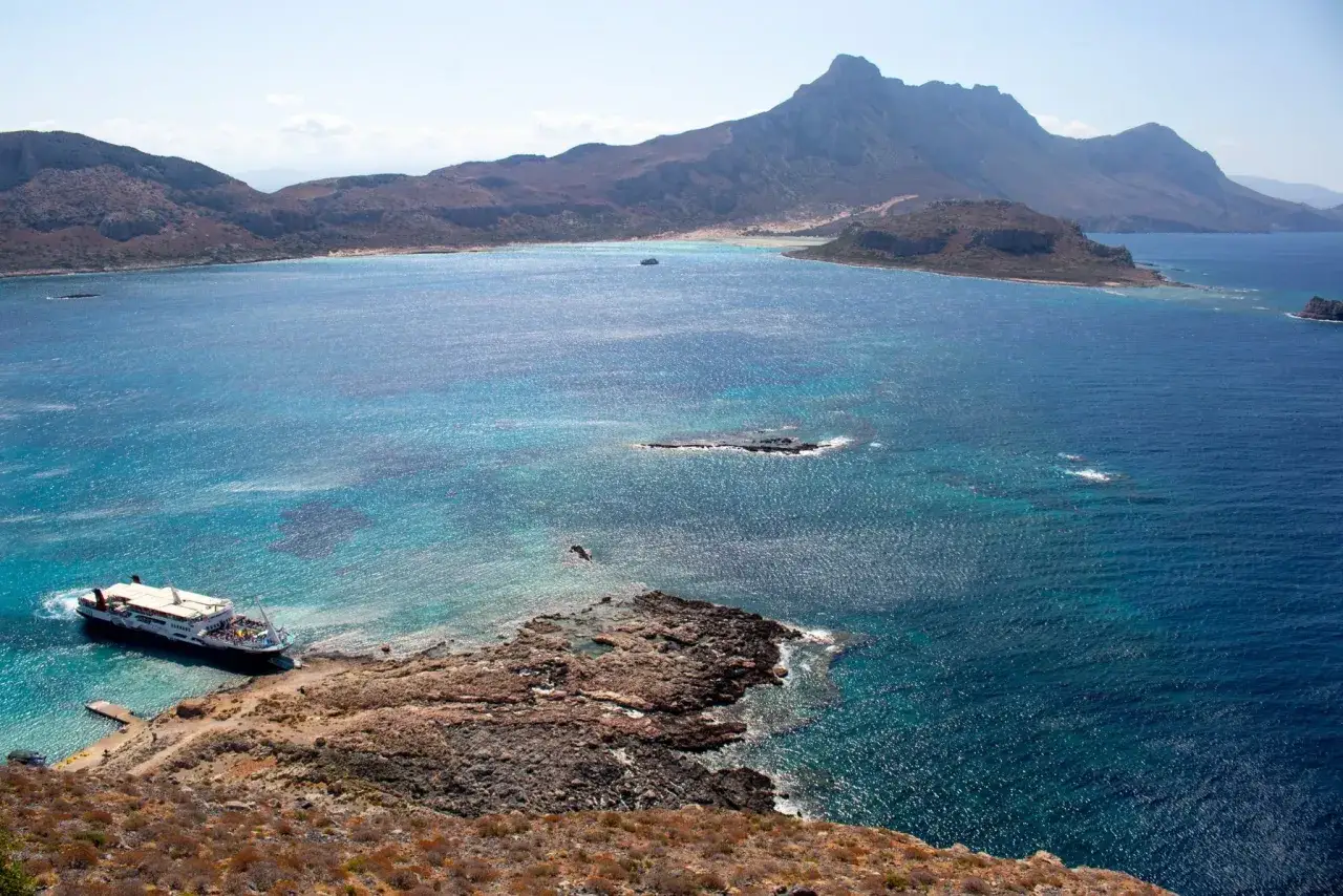 Statek przycumowany do skalistego brzegu, turkusowa woda, góry w tle. Czy to Korfu czy Kreta?