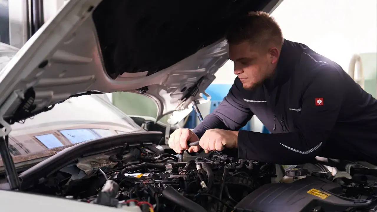 Mechaniker mit Bart arbeitet konzentriert am Motor eines Autos in einer **freien Werkstatt**.