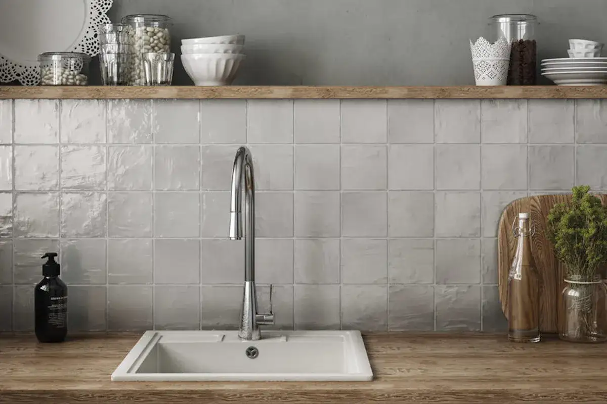 Crédence de cuisine avec carrelage gris clair et joints discrets. Un évier blanc et un robinet chromé sont visibles, ainsi que des bocaux sur une étagère en bois.