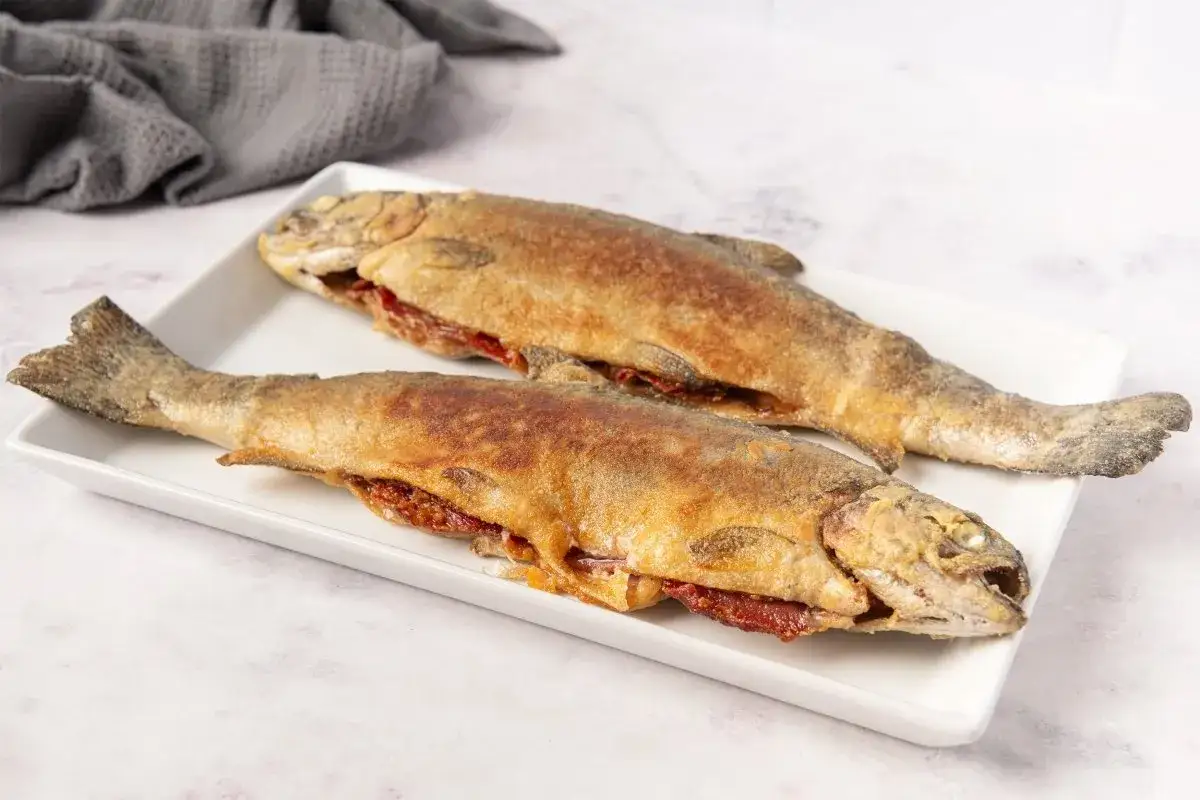 Dos truchas a la navarra doradas y crujientes, rellenas de pimientos rojos, servidas en una bandeja blanca.