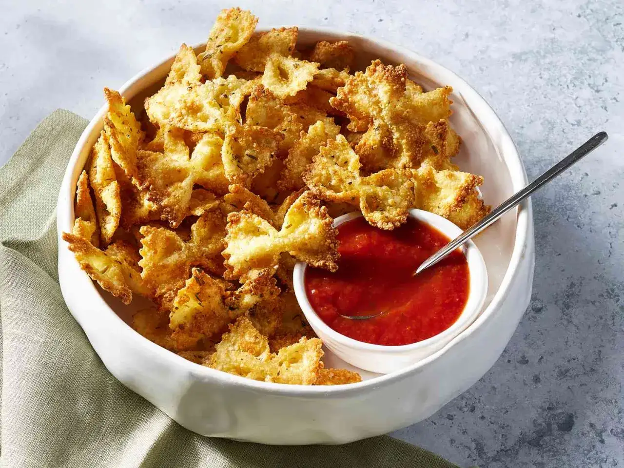 Krispiga pasta chips i form av fjärilar, serverade i en vit skål med en liten skål tomatsås och en sked.
