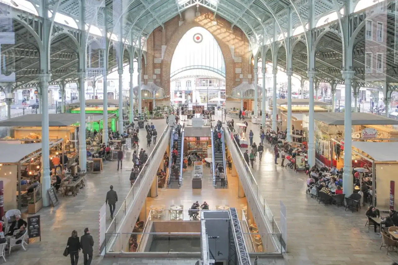 Interior del mercado de Colón, con gente comprando y comiendo en puestos y restaurantes bajo una estructura metálica y arcos de ladrillo.