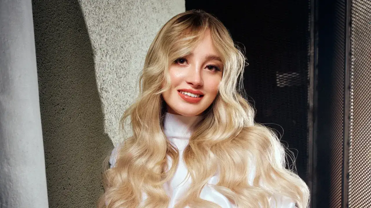 Ragazza con lunghi capelli biondi ondulati, indossa un abito bianco a collo alto. Il suo sorriso &egrave; radioso, con labbra color **pantone crema**.