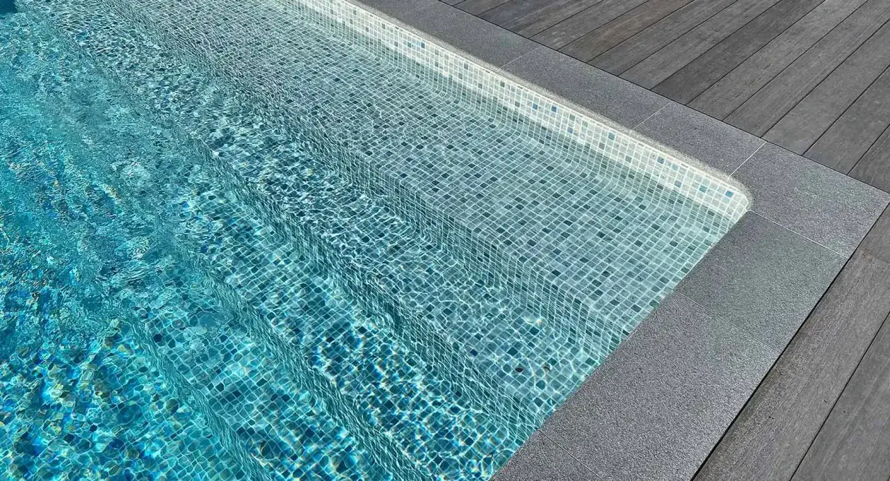 Trappsteg i en pool med bl&aring; mosaik, kantade av gr&aring; sten och ett tr&auml;d&auml;ck. En inbjudande sarg runt poolen.