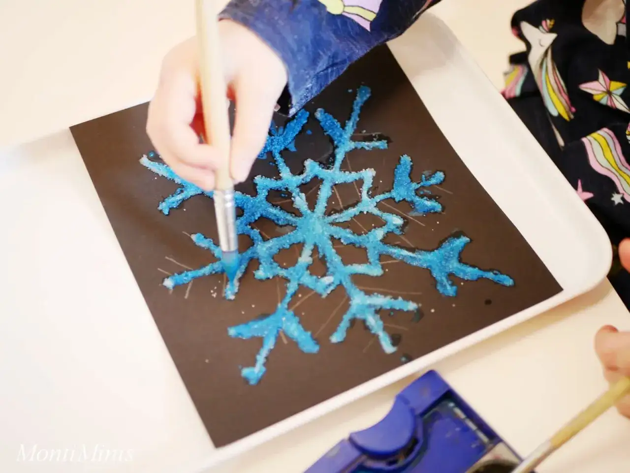 Kind malt mit Pinsel und blauer Farbe auf schwarzem Papier eine Schneeflocke, die mit Salz bedeckt ist. Tolle Salzbilder!
