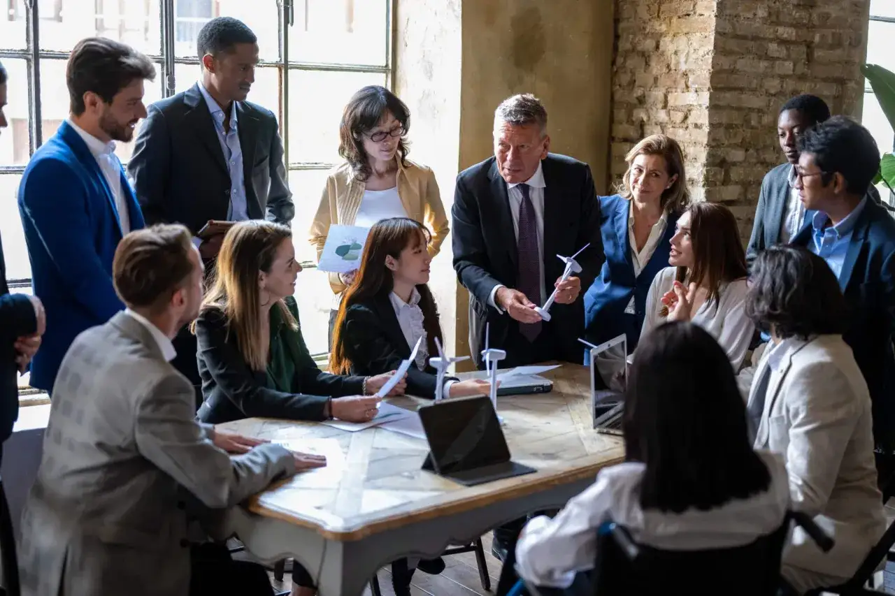 A diverse team discusses a project, with a man holding a wind turbine model. This scene offers insights on how to ask for promotion by showcasing collaboration and strategic thinking.