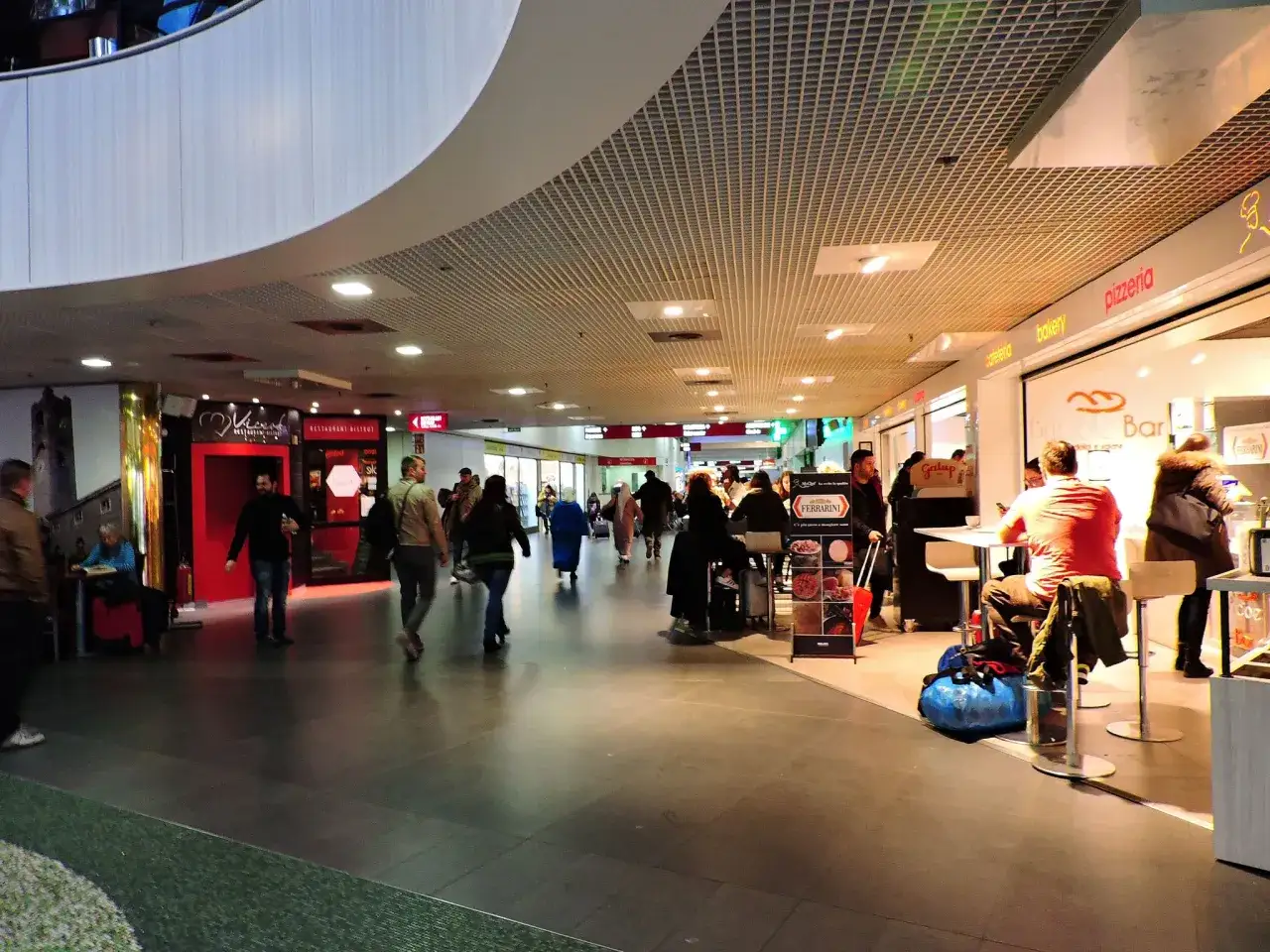 Persone passeggiano nel corridoio di un aeroporto, con negozi come pizzeria e bakery. Un uomo &egrave; seduto a un tavolo. Milano Bergamo aeroporto.