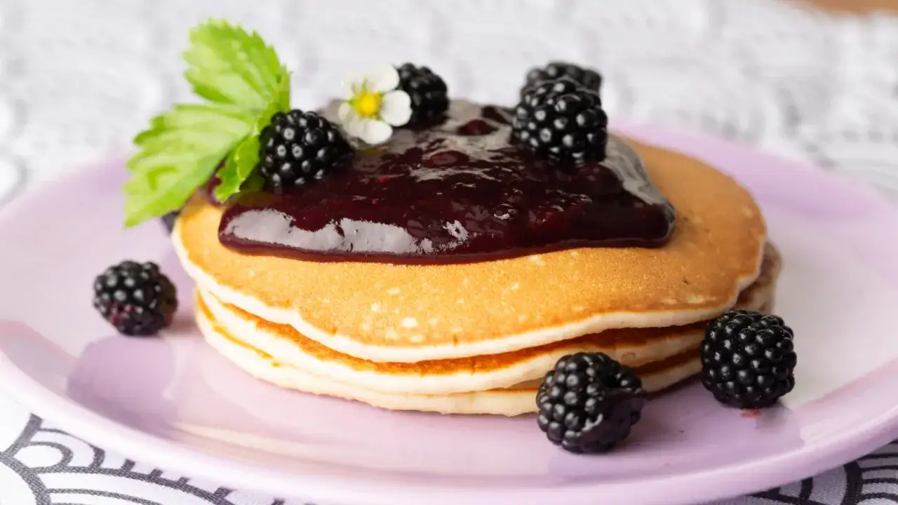 Pancake senza uova con marmellata di more e frutti di bosco freschi, guarniti con una fogliolina verde e un fiorellino bianco.