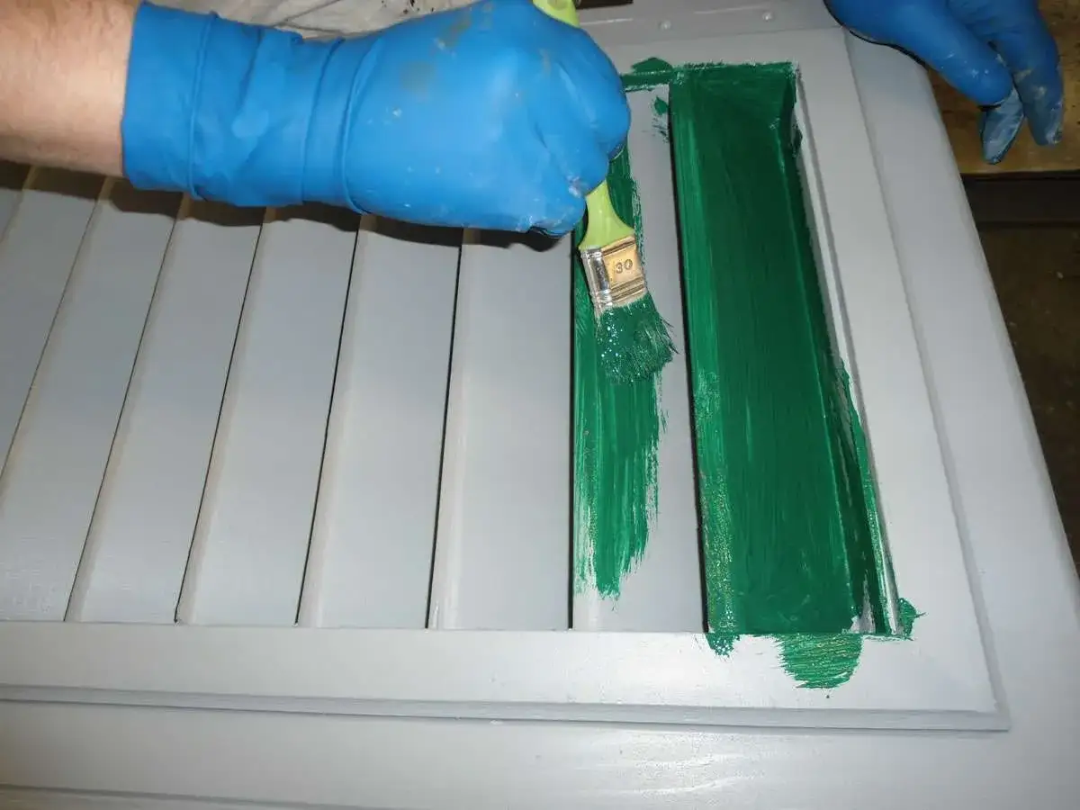Mano con guanto blu che dipinge con pennello verde una persiana grigia. La verniciatura persiane è in corso.