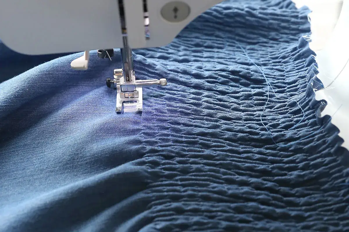Sewing machine creating elastic smocking on blue fabric. The fabric is gathered into neat rows, giving it a textured, ruched appearance.