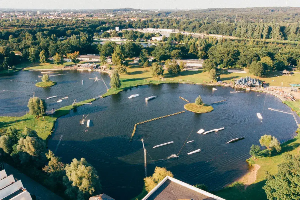 Luftaufnahme vom Strandbad Wedau mit Wakeboard-Anlage, Sprungschanzen und gr&uuml;nen Ufern.
