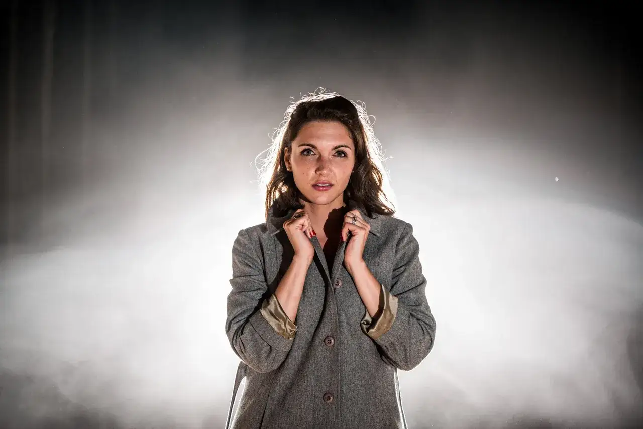 Une jeune femme au regard intense, photo au flash, ajuste le col de son manteau gris. La lumière met en valeur ses cheveux et son visage.