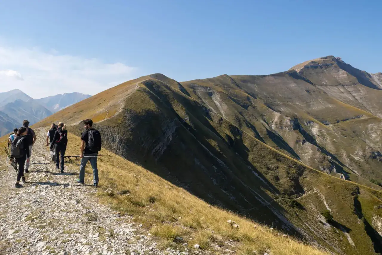 Escursionisti sul sentiero che porta alla grotta della Sibilla, tra montagne verdi e rocciose sotto un cielo azzurro.