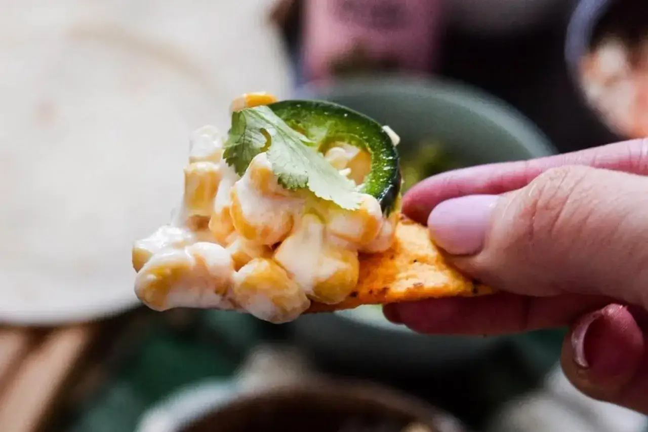 En hand håller en nachochips med krämig majs, jalapeño och koriander.