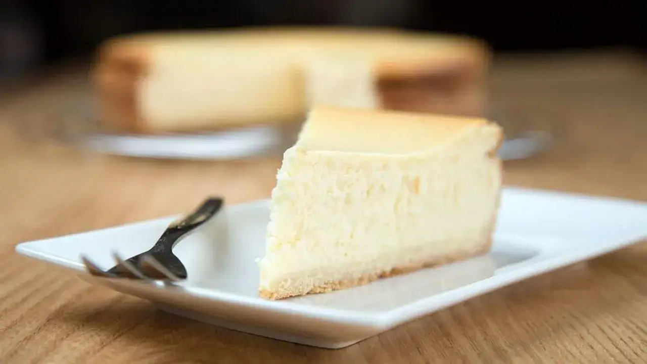 Ein Stück cremiger Quarkkuchen ohne Boden auf einem weißen Teller, daneben eine Gabel. Im Hintergrund ist der ganze Kuchen zu sehen.