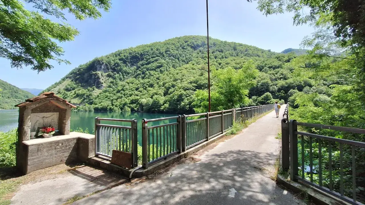 Un piccolo edicola votiva con fiori, un ponte con ringhiera e una persona che cammina, con montagne verdi e un lago sullo sfondo. I **vagli di sotto** offrono una vista mozzafiato.
