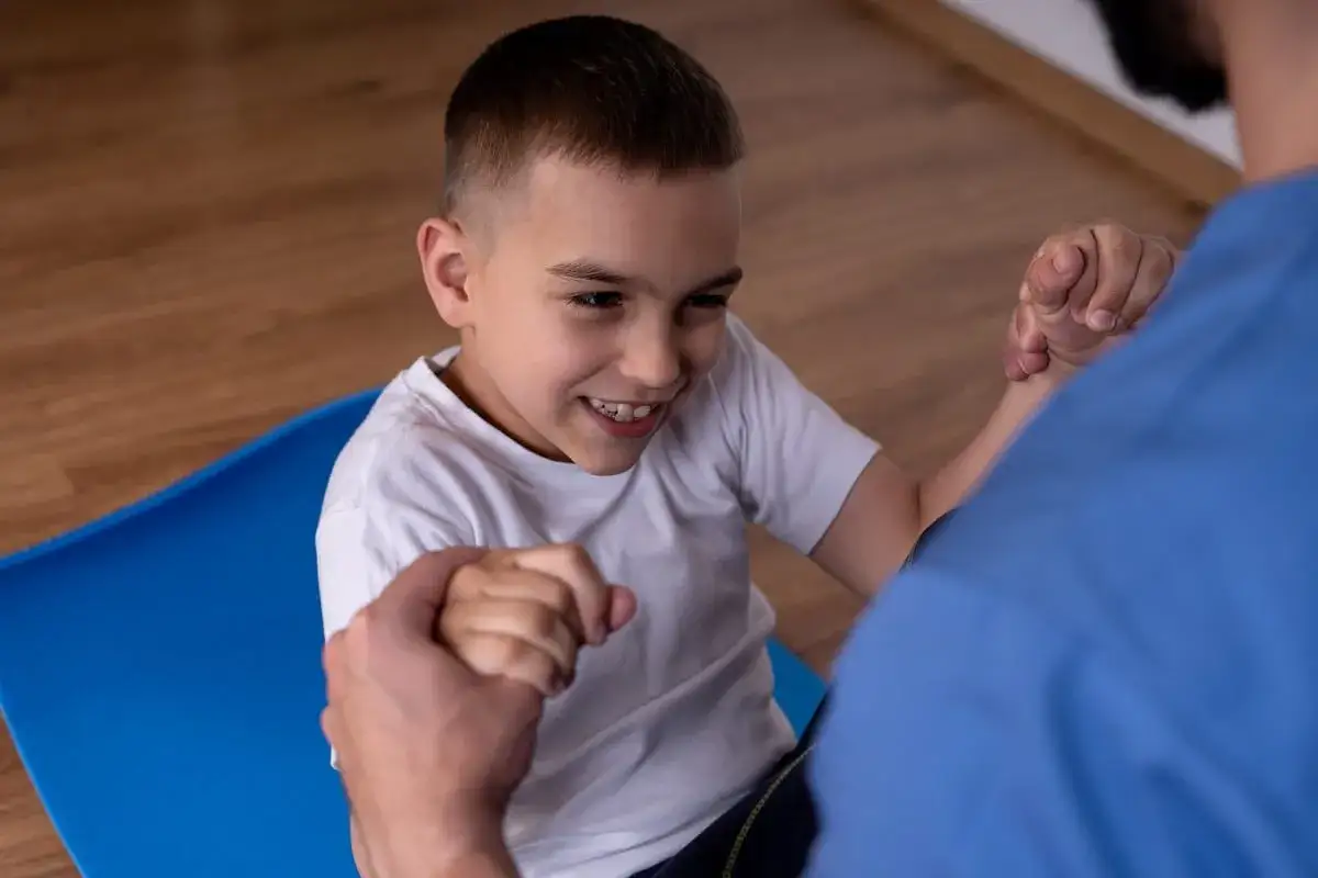 Ni&ntilde;o sonriente en fisioterapia, trabajando en su movilidad con ayuda. Su **par&aacute;lisis cerebral esp&aacute;stica** no le impide progresar.