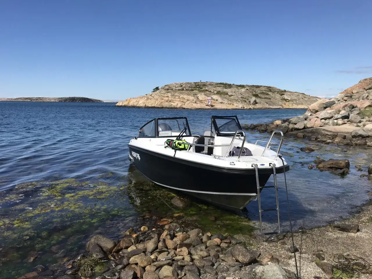 En svartvit b&aring;t ligger vid en stenig strand i Grebbestad fjorden. Klart bl&aring; himmel och lugnt vatten.