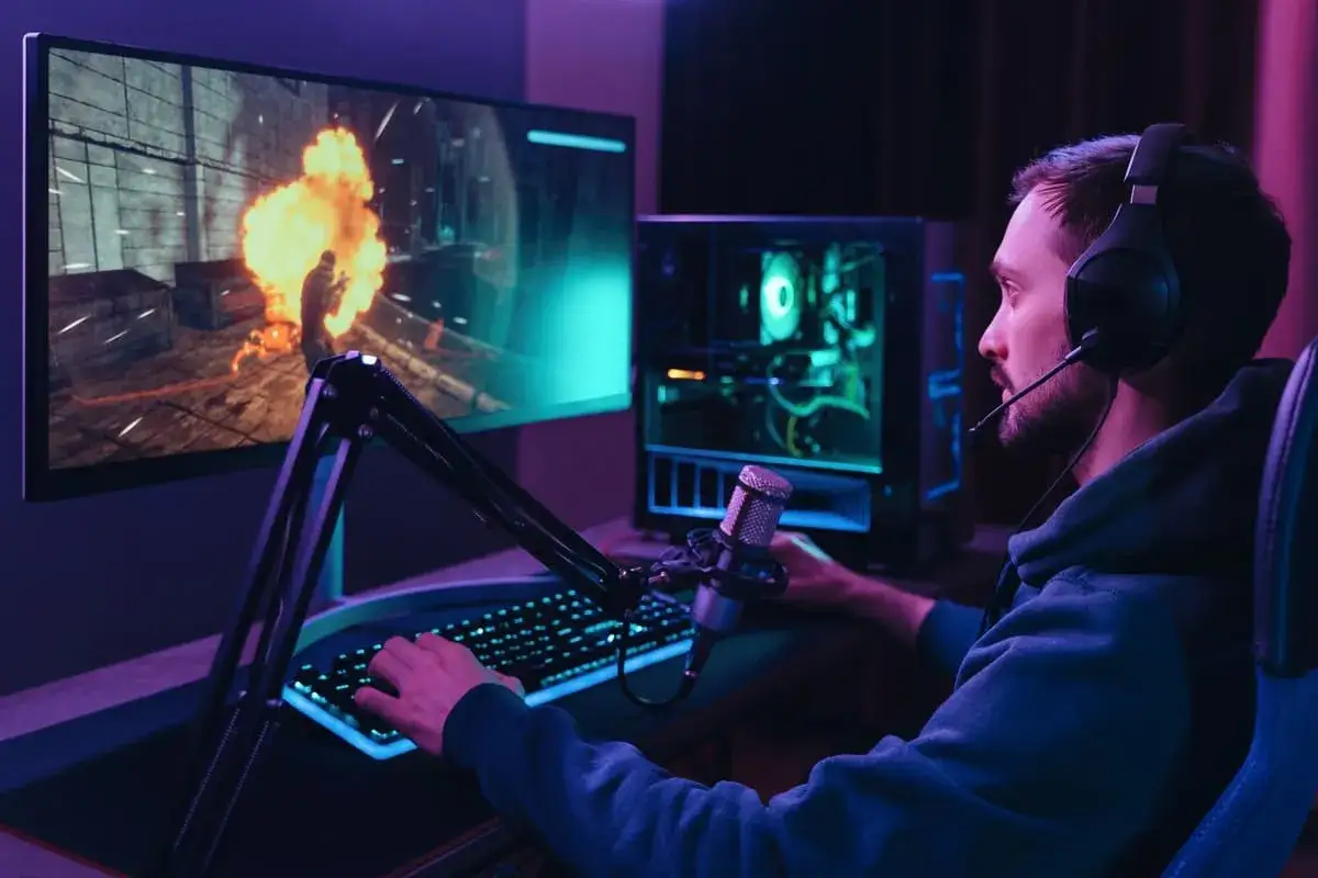 Jugador con auriculares y micr&oacute;fono, rodeado de perif&eacute;ricos de salida como monitor y PC iluminados, inmerso en un juego de acci&oacute;n.