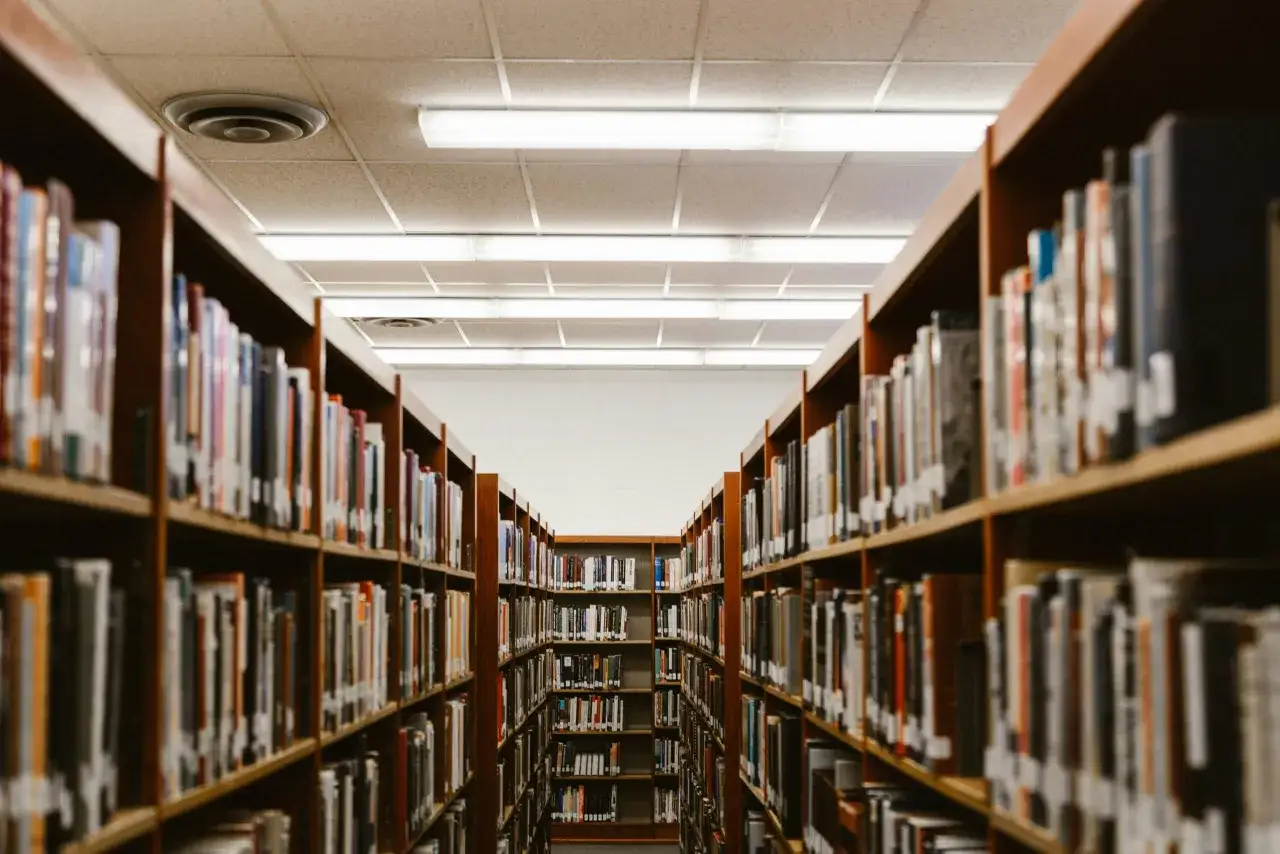 Wąskie przejście między regałami pełnymi książek w bibliotece. W tle widać rzędy oświetlenia sufitowego, idealne do nauki i rozwijania **fce use of english**.